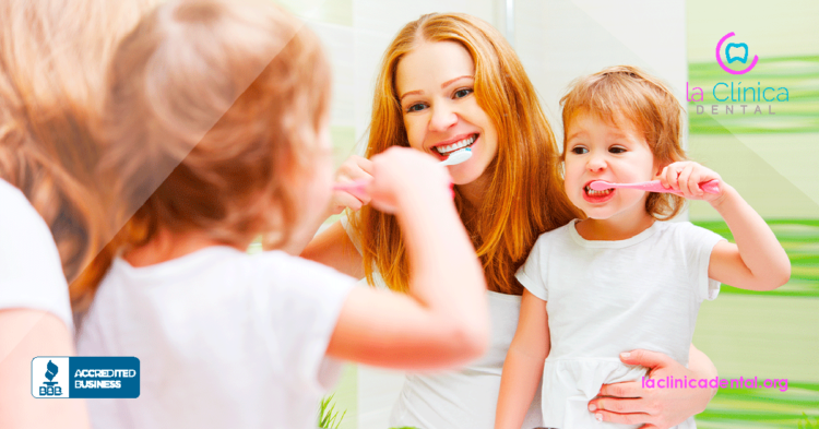 Mujer y niña cepillándose los dientes en un baño, promoviendo la higiene dental infantil, con el logo de La Clínica Dental Guadalajara en la esquina superior derecha.