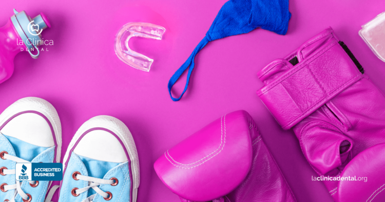 Zapatillas deportivas, protector bucal y guantes de boxeo sobre fondo rosa, representando la importancia de la protección dental en deportes, relacionado con la clínica dental.