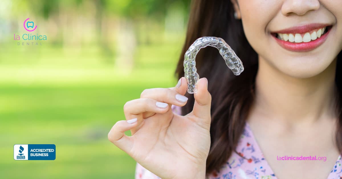 Mujer sonriente sosteniendo alineador dental transparente de Invisalign en un entorno natural, representando tratamientos estéticos en La Clínica Dental Guadalajara.
