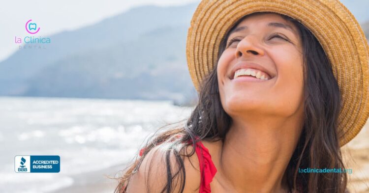 Mujer sonriendo en la playa, usando sombrero, destacando la importancia de la salud dental durante las vacaciones, con logo de La Clínica Dental Guadalajara.