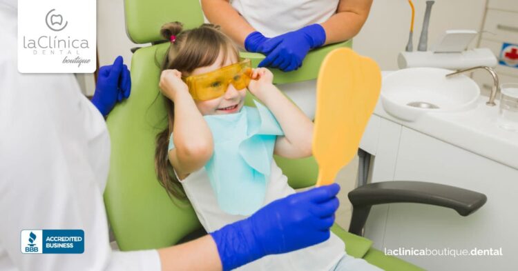 Niña sonriente en silla dental con gafas de protección, sosteniendo un espejo, en La Clínica Dental Guadalajara, enfocada en cuidado dental infantil.