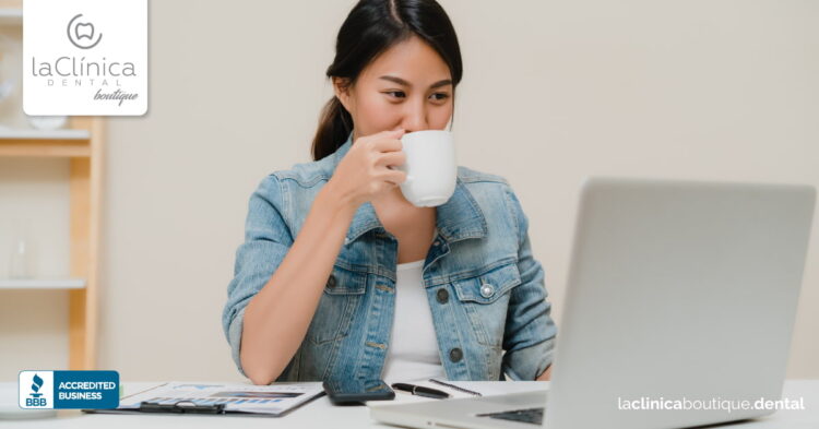 Mujer sosteniendo una taza, mirando una computadora portátil en la clínica dental, simbolizando atención al paciente y servicios de salud dental.